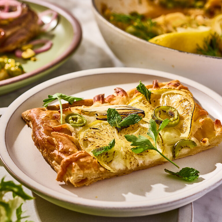 Sliced flatbread on a white plate topped with squash, jalapenos, mint and parsley leaves