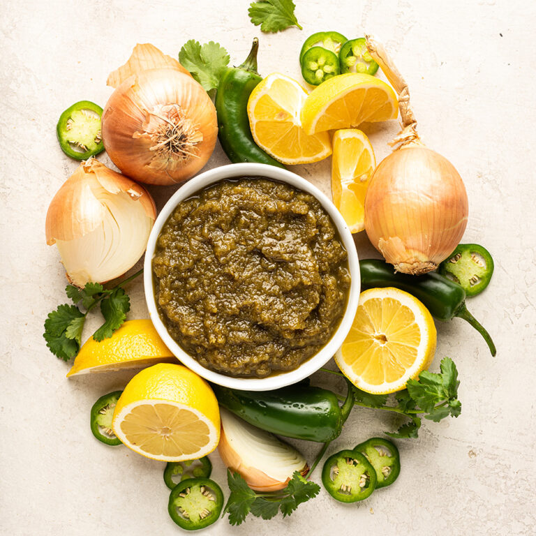 A bowl of chutney surrounded by whole white onions, lemon slices, whole and sliced jalapeno