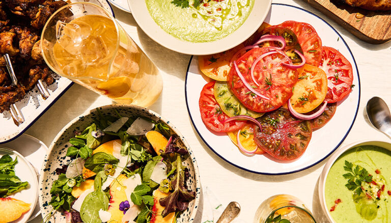 A spread of summer food such as iced tea, sliced tomatoes, peach salad, and chicken skewers